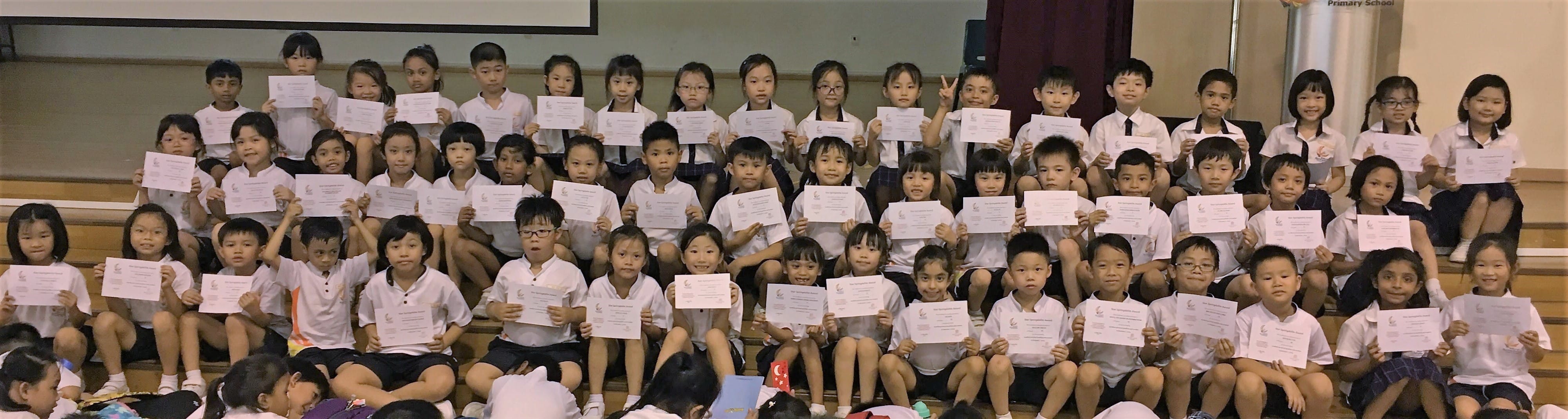 Large group of young students in white uniforms sitting on bleachers, each holding a certificate.